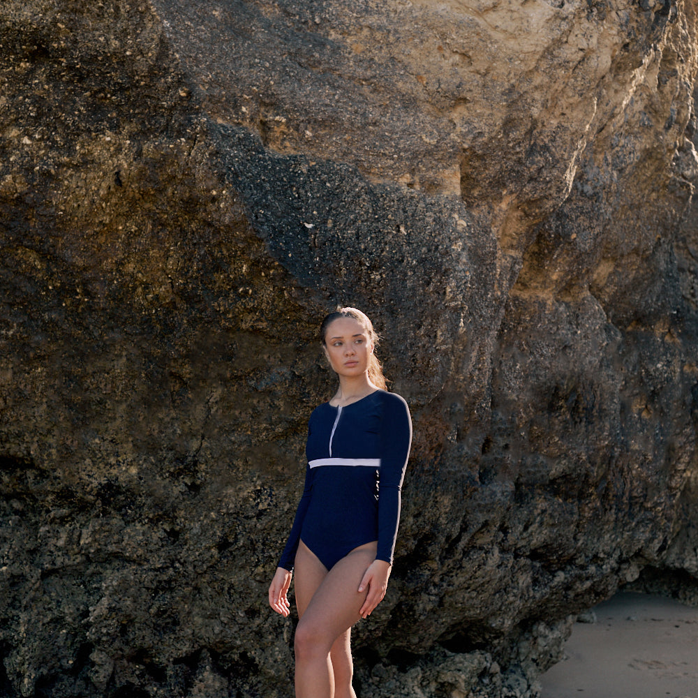Woman wearing a marine blue swimsuit with long sleeves, white button strip and white line under the bust, in front of a rocky background.