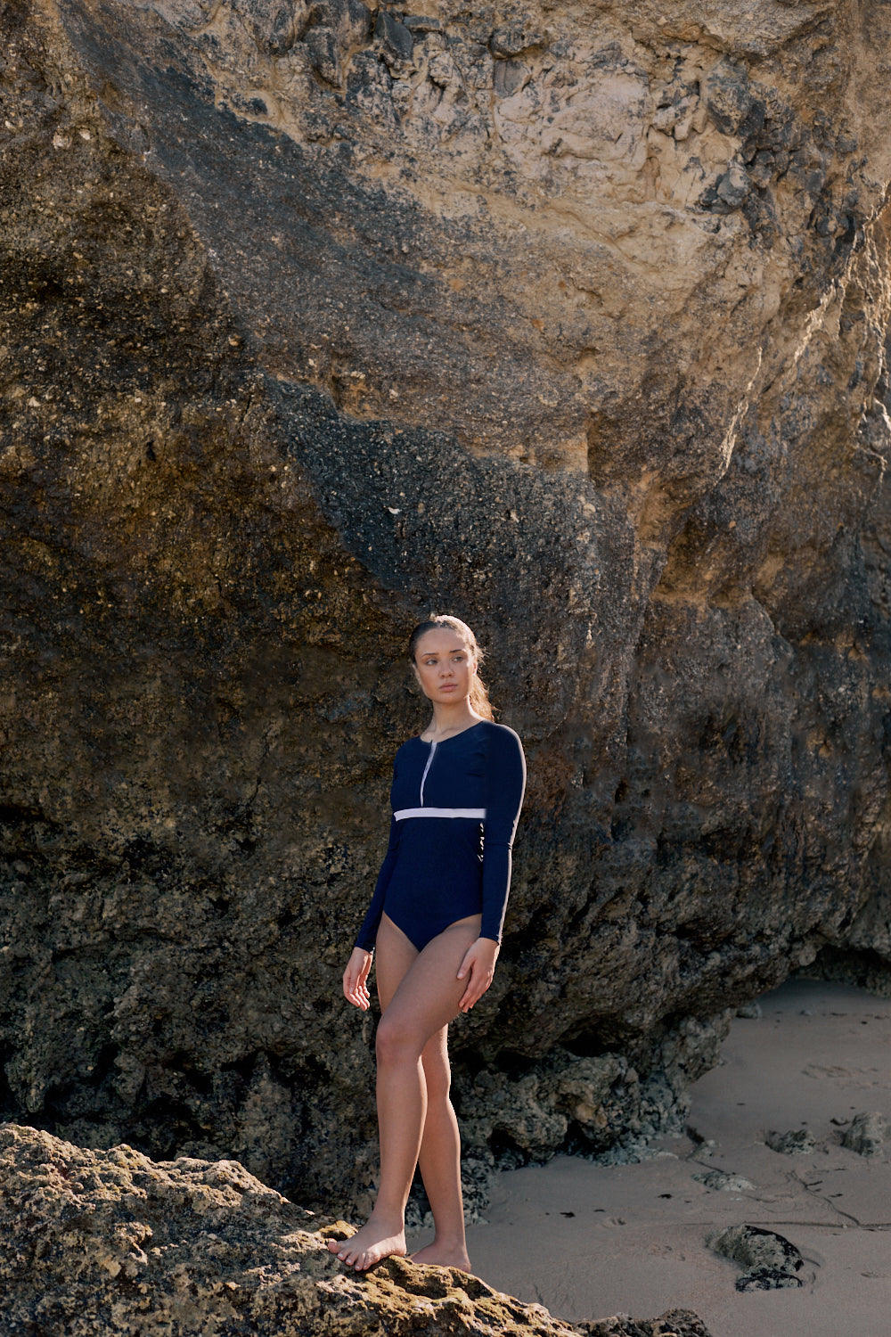 Woman wearing a marine blue swimsuit with long sleeves, white button strip and white line under the bust, in front of a rocky background.