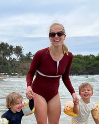 Woman in a red swimsuit with long sleeves with two children on a beach.