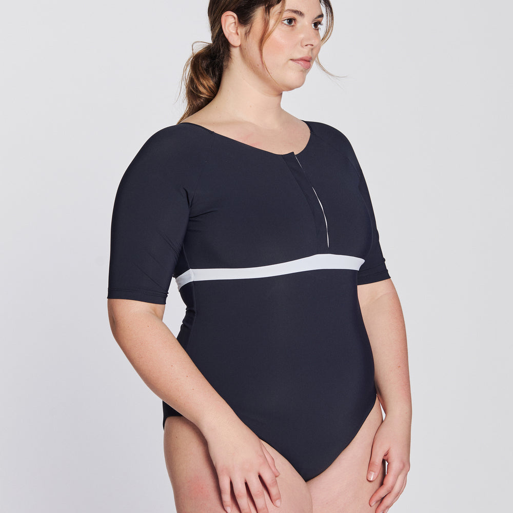 
                  
                    Side view of a woman wearing a marine blue swimsuit with short sleeves, white button strip and white line under the bust, in front of a white background.
                  
                