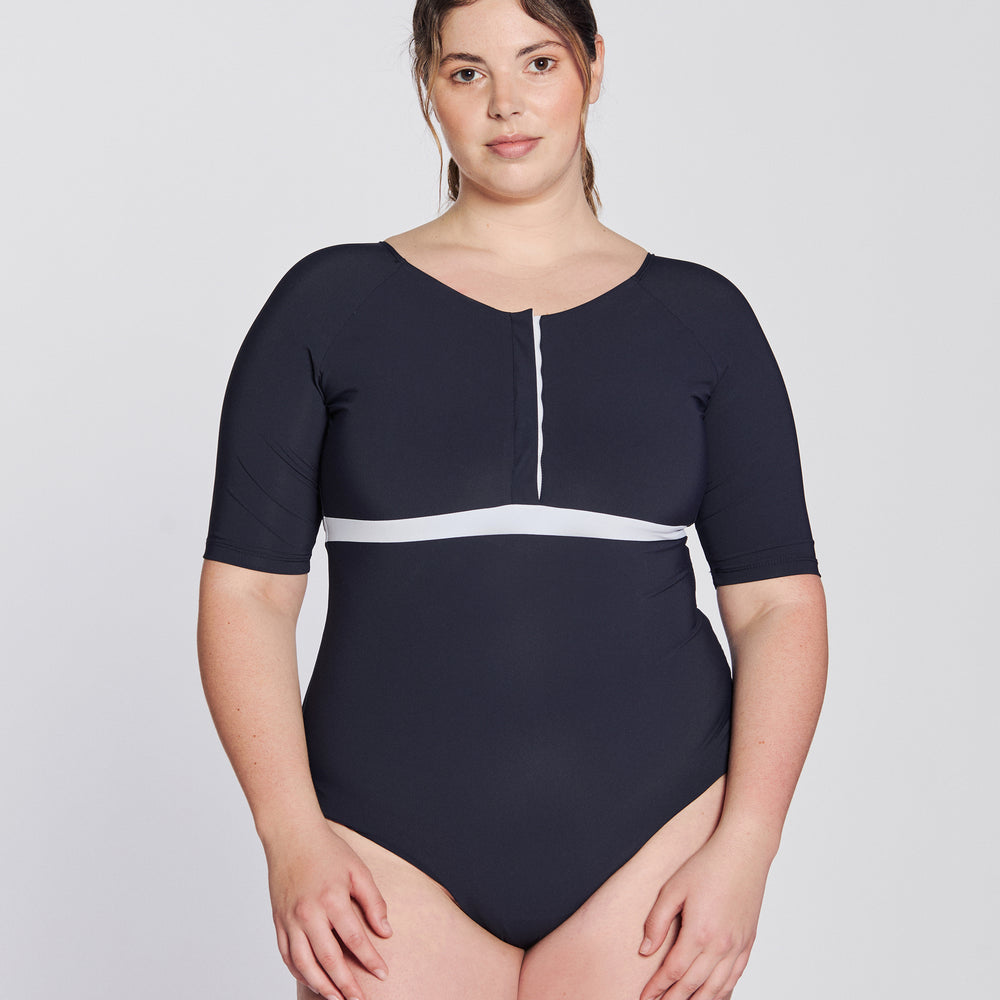 Front view of a woman wearing a marine blue swimsuit with short sleeves, white button strip and white line under the bust, in front of a white background.