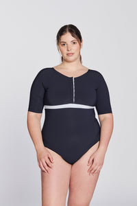 Front view of a woman wearing a marine blue swimsuit with short sleeves, white button strip and white line under the bust, in front of a white background.