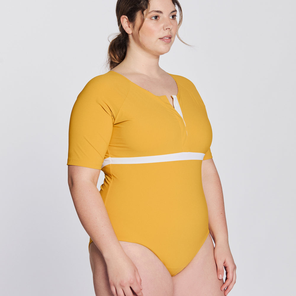 
                  
                    Side view of a woman wearing a sunflower yellow swimsuit with short sleeves, white button strip and white line under the bust, in front of a white background.
                  
                