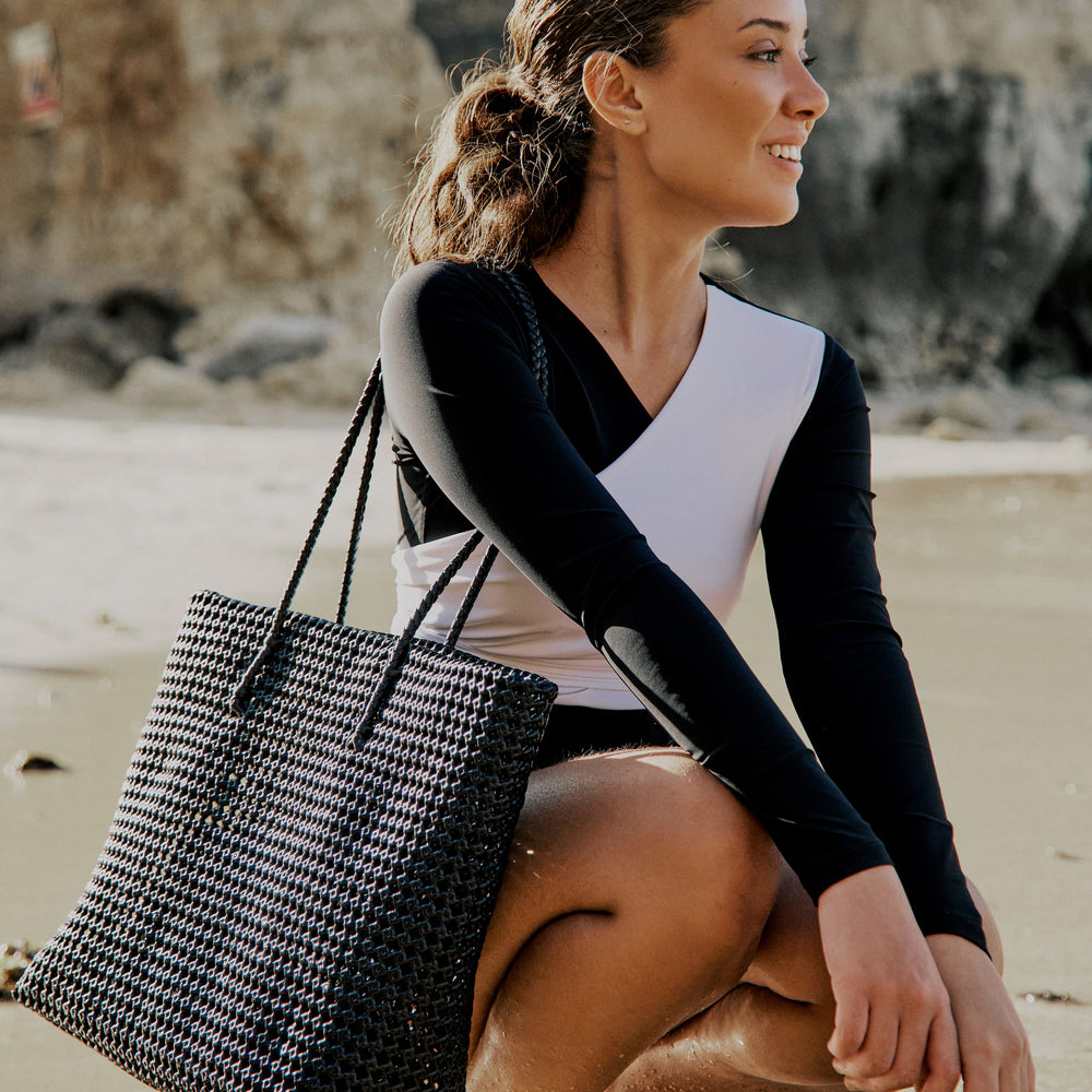 Side view of a woman wearing a black and white asymmetrical swimsuit with long sleeves, on a beach.