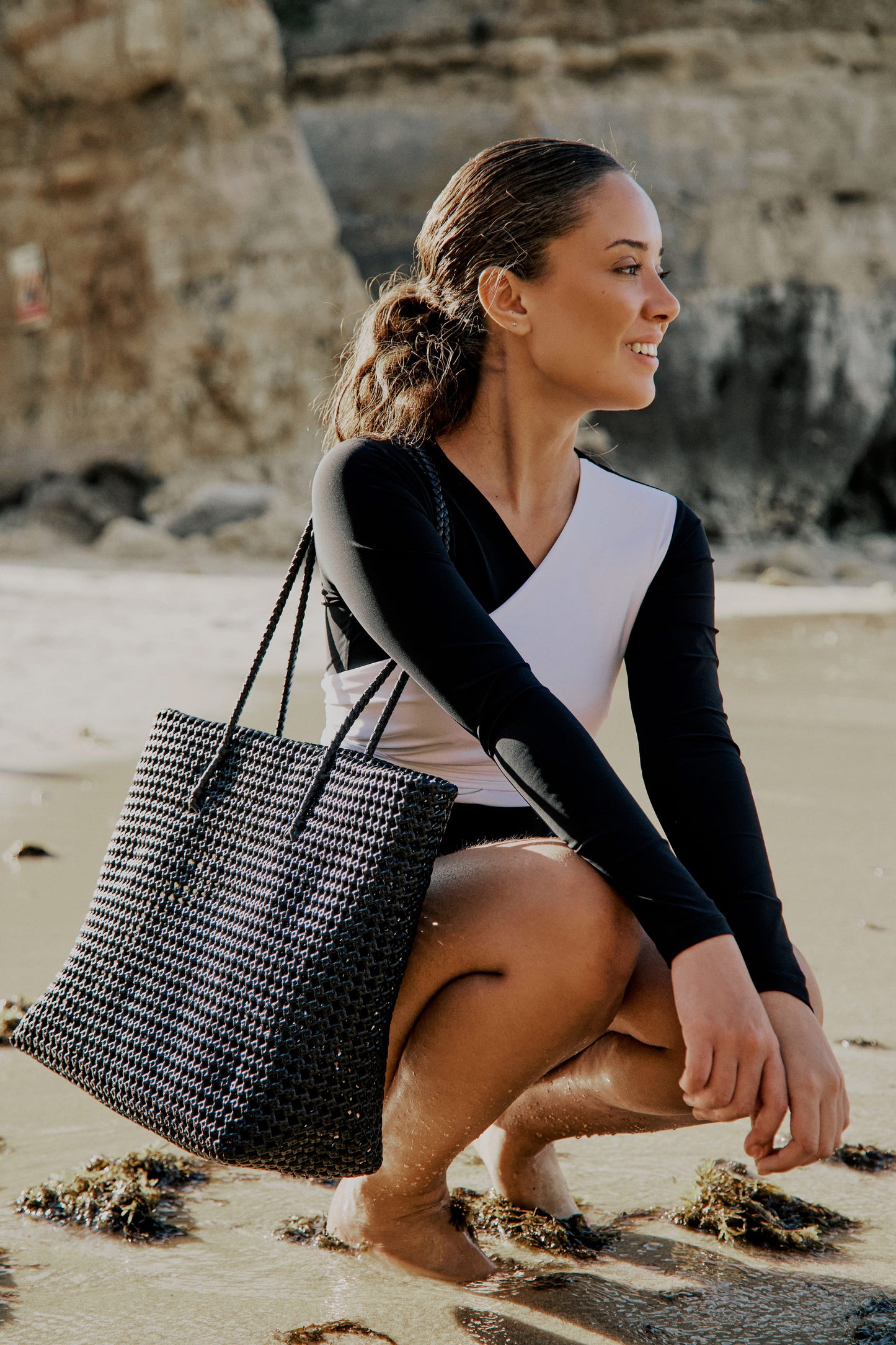 Side view of a woman wearing a black and white asymmetrical swimsuit with long sleeves, on a beach.