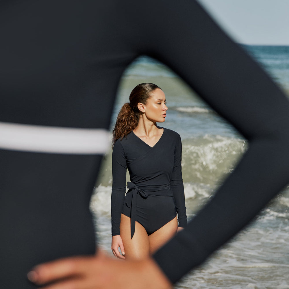 Woman wearing a marine blue swimsuit with long sleeves and wrap closing, in front of the sea.
