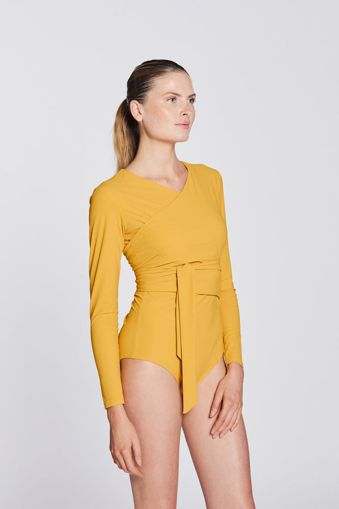 
                  
                    Side view of a woman wearing a sunflower yellow swimsuit with long sleeves and wrap closing, in front of a white background.
                  
                
