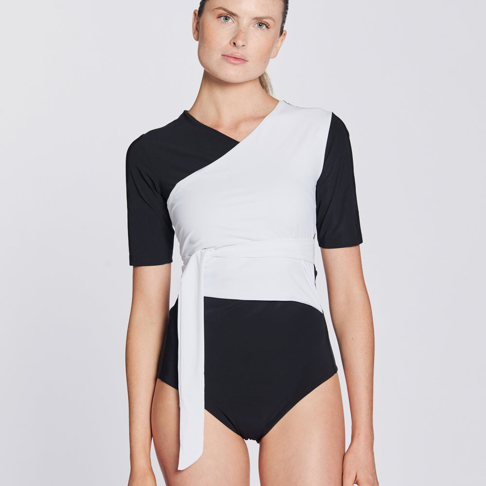 Front view of a woman wearing a black and white asymmetrical swimsuit with short sleeves, on a white background.