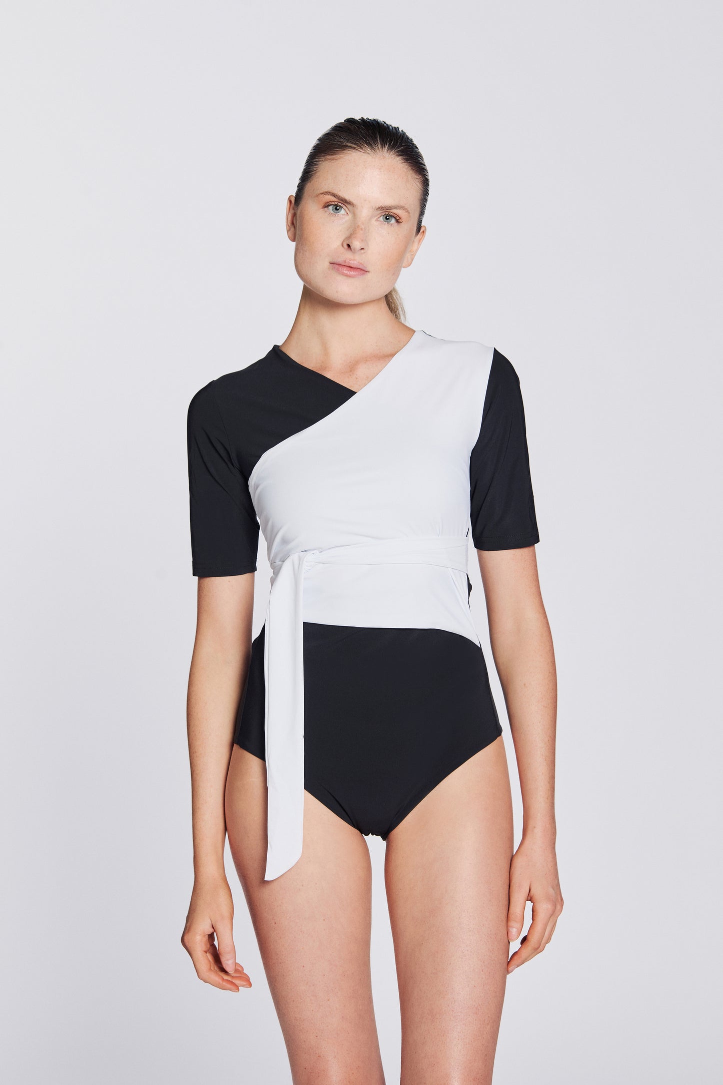 Front view of a woman wearing a black and white asymmetrical swimsuit with short sleeves, on a white background.