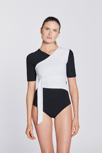 Front view of a woman wearing a black and white asymmetrical swimsuit with short sleeves, on a white background.