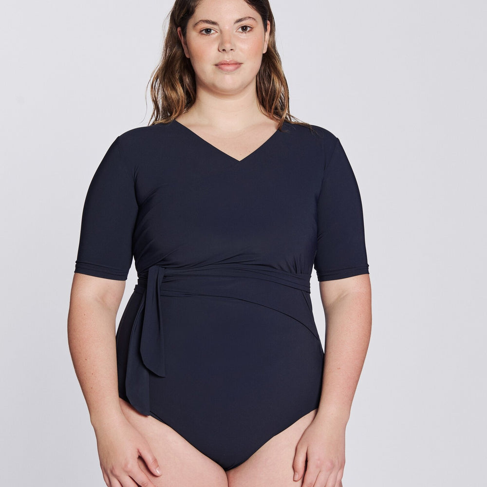 
                  
                    Front view of a woman wearing a marine blue swimsuit with short sleeves and wrap closing, in front of a white background.
                  
                