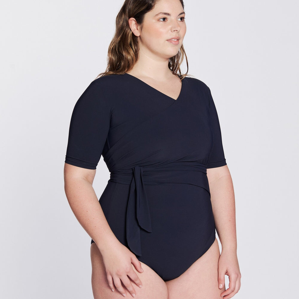 
                  
                    Side view of a woman wearing a marine blue swimsuit with short sleeves and wrap closing, in front of a white background.
                  
                