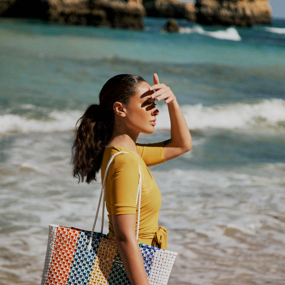 
                  
                    Woman with a colorful beach bag standing on a beach with rocky cliffs and ocean waves in the background.
                  
                